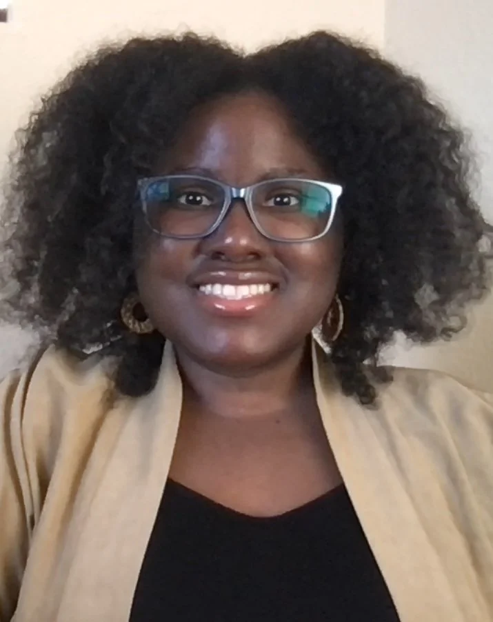 A woman with dark skin and curly black hair, wearing glasses, gold earrings, a black top, and a beige jacket, smiling at the camera.