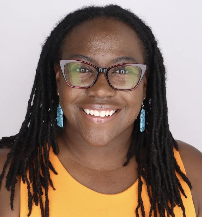 A smiling woman with glasses and blue earrings, wearing an orange top, against a plain light background.