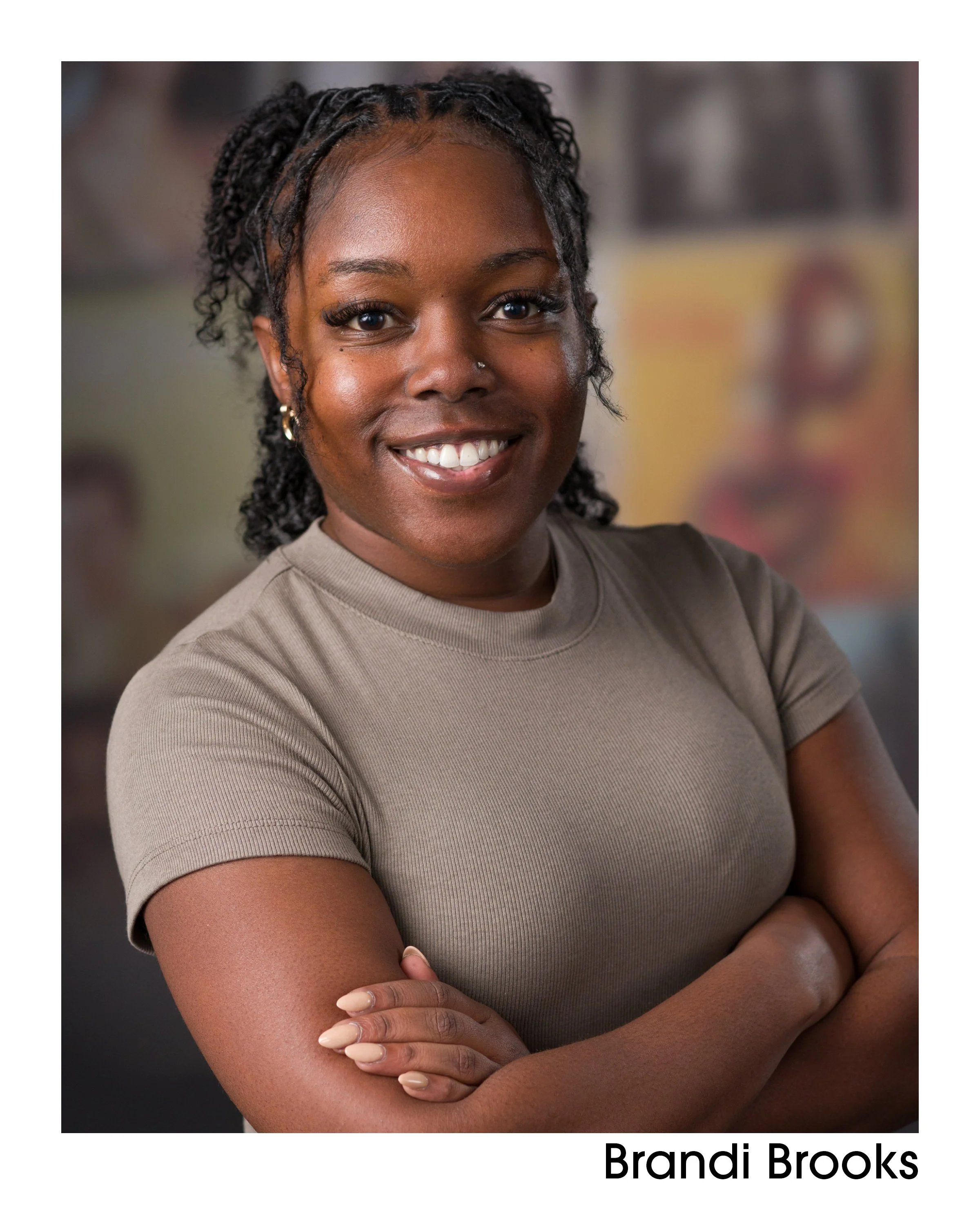 A woman with dark skin and curly hair styled in twists, smiling with arms crossed, wearing a beige t-shirt, with a blurred background of colorful artwork.