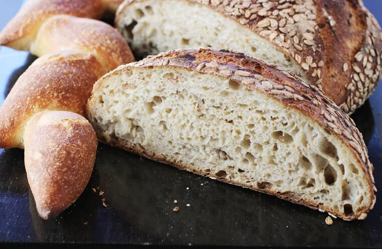 Close-up of a sliced loaf of rustic bread with a golden crust and airy interior, accompanied by smaller bread rolls, all placed on a dark surface.