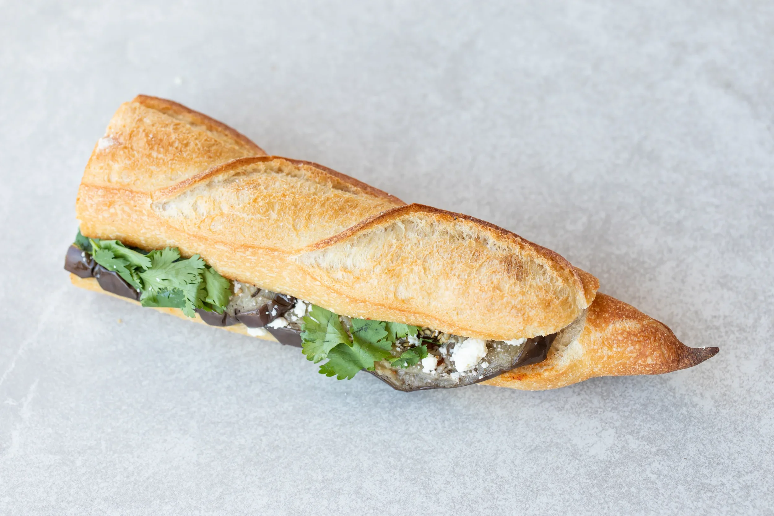 A baguette sandwich with dark lettuce, crumbled white cheese, and fresh cilantro