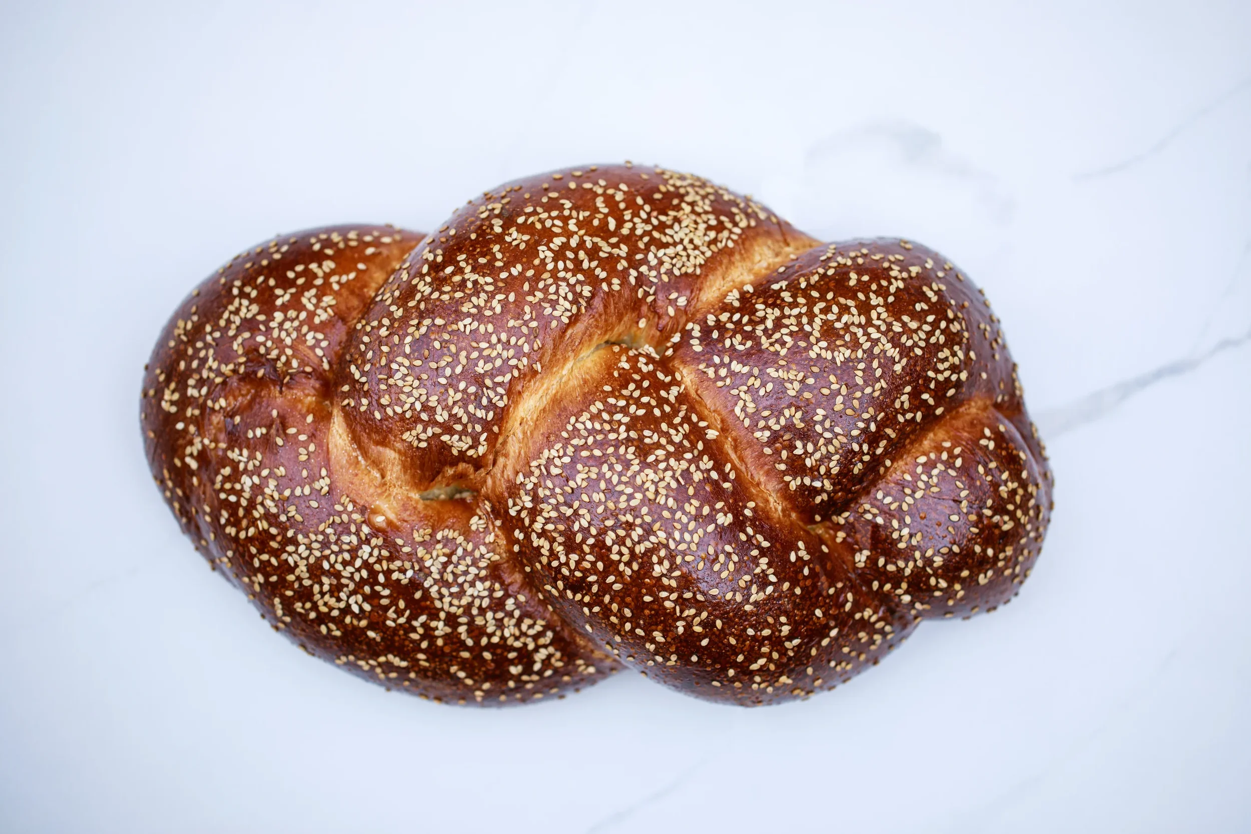 A braided challah bread loaf topped with sesame seeds on a white marble surface.