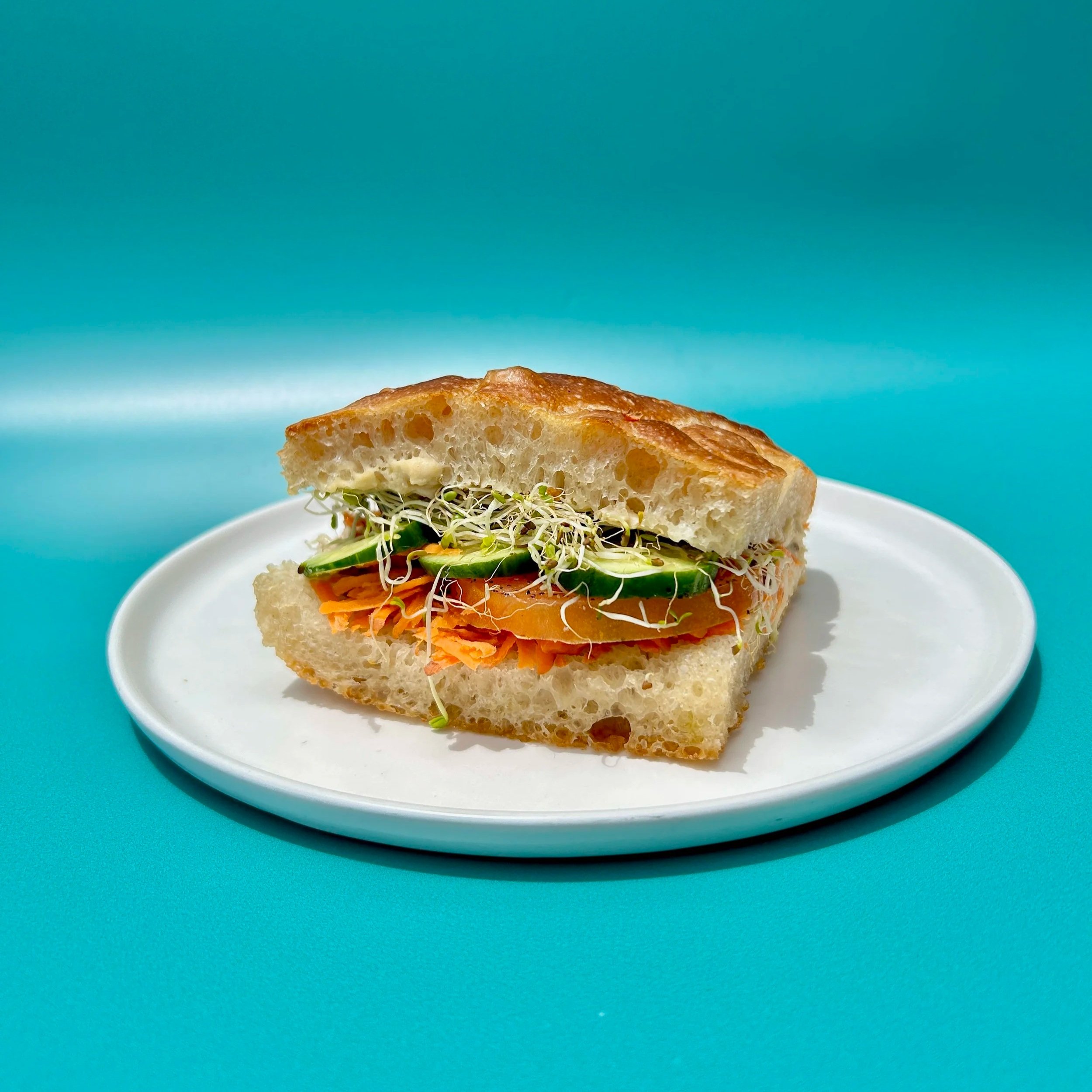 We spread garlicky hummus on our focaccia bread. We then stack crunchy shredded carrots, sliced cucumbers tossed in lemon juice, fresh slices of tomato, and alfalfa sprouts.