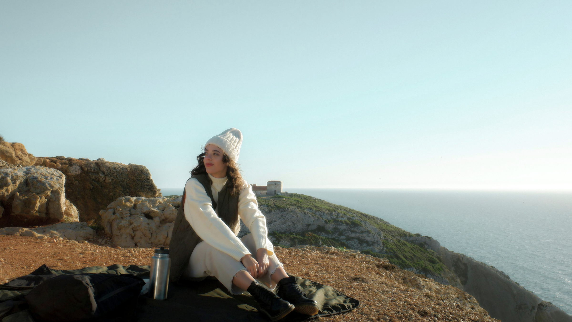 A young woman sitting on a blanket on a rocky hillside overlooking the ocean, with a historic building visible in the distance under a clear sky.