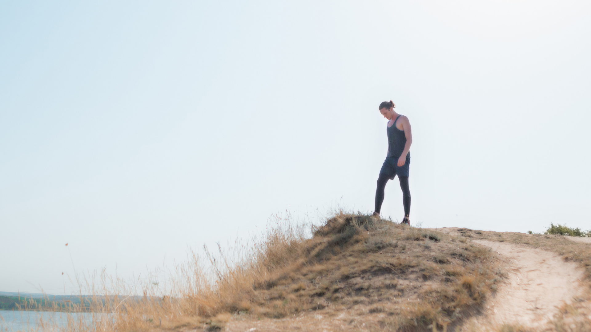 A woman standing on a small hilltop outdoors, looking down, with a path nearby and dry grass around, under a clear sky.