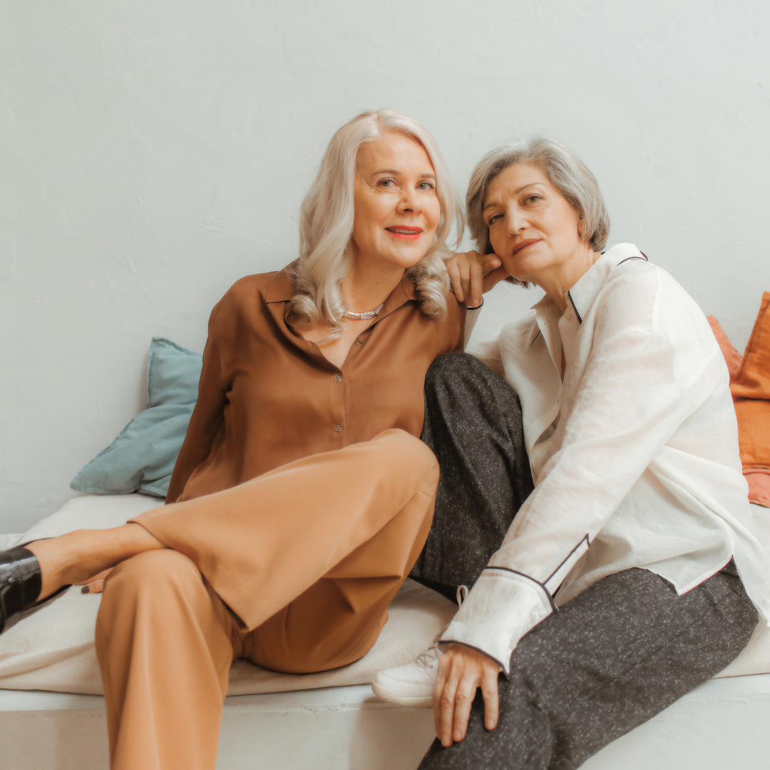 Two mature women sitting close together on a bed, both with silver hair, one in brown pajamas and the other in light-colored pajamas, looking at the camera with relaxed expressions.