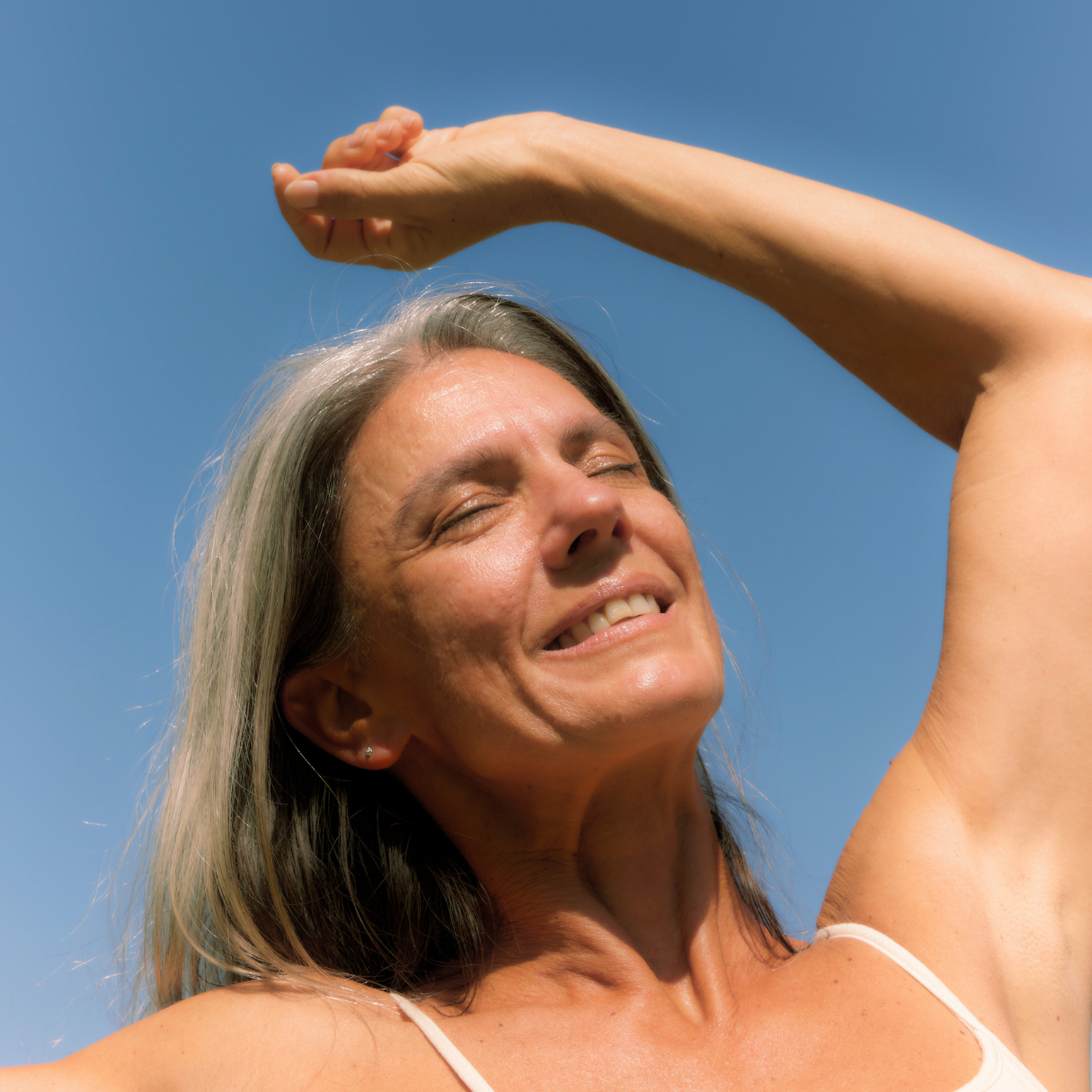 A woman smiling with eyes closed, enjoying sunshine outdoors, with her hand resting on her forehead.