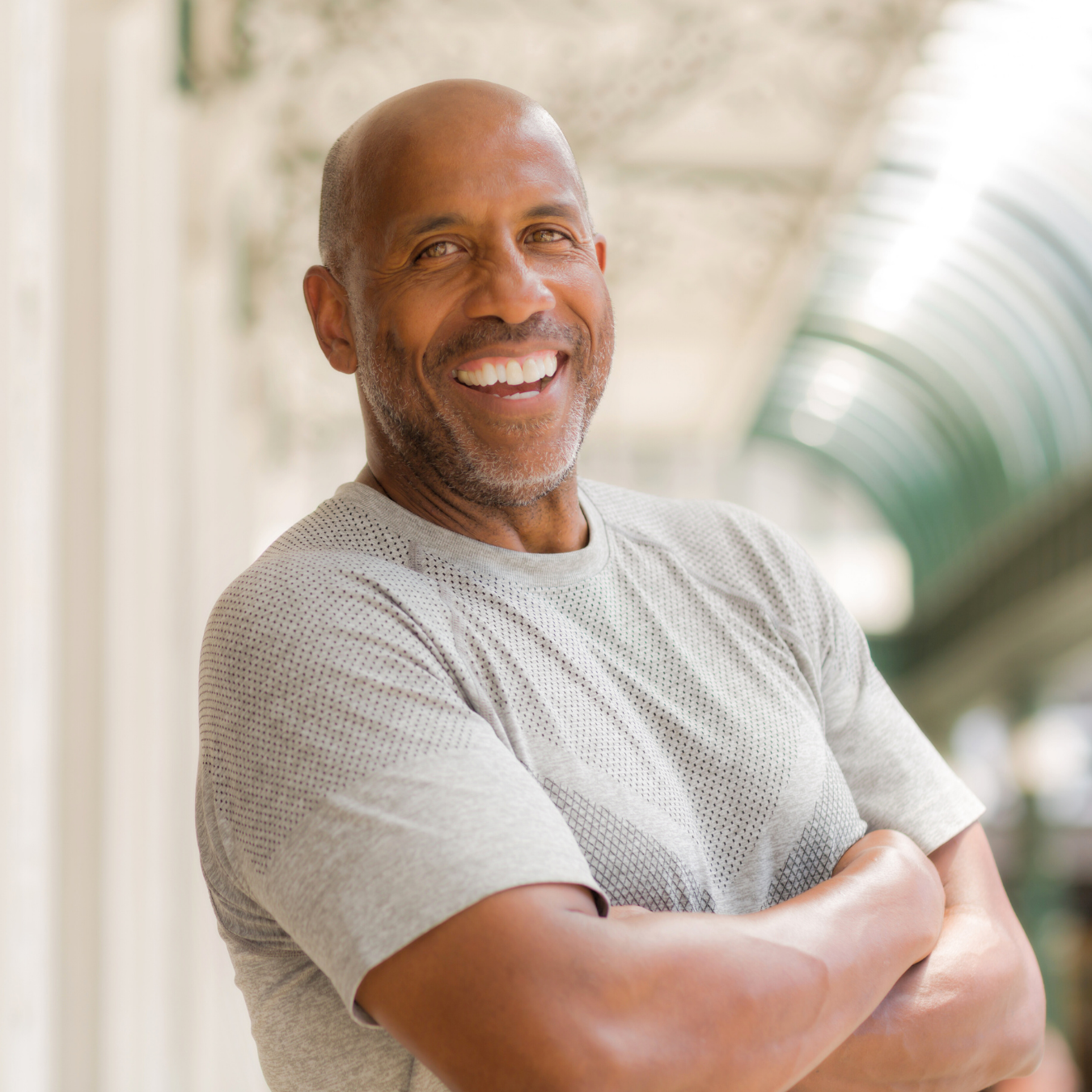 A smiling middle-aged man with a beard and bald head, wearing a light gray t-shirt with his arms crossed, standing in a bright, airy space with a high, arched, glass ceiling.
