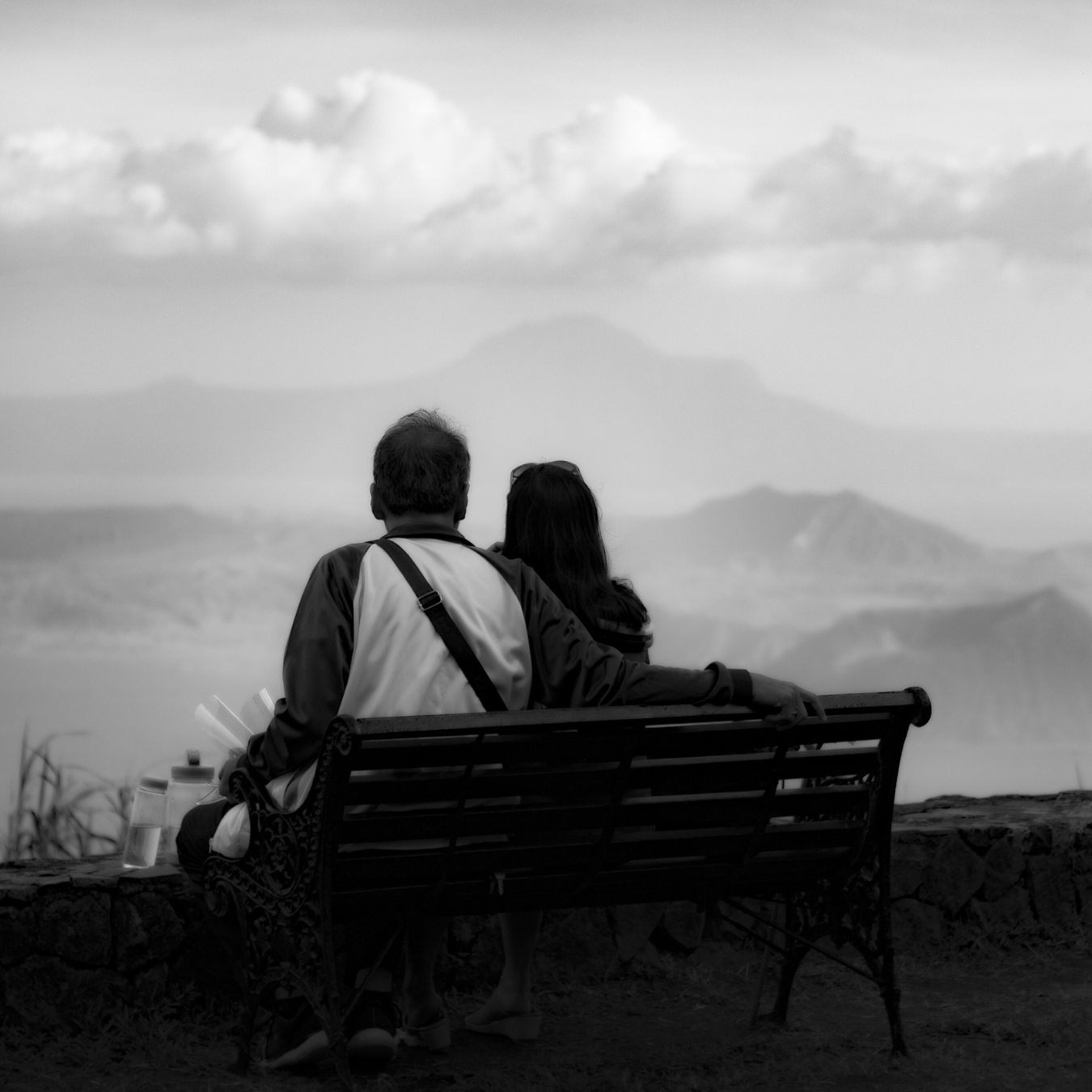 A black and white photo of a man and woman sitting on a park bench, overlooking a mountainous landscape with clouds in the sky.