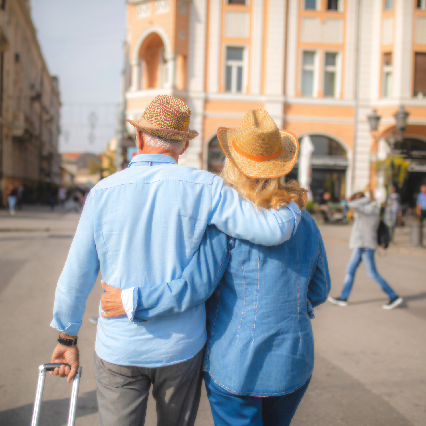 Older couple walking arm in arm through a city street, both wearing straw hats and casual clothing, with buildings and other pedestrians in the background.