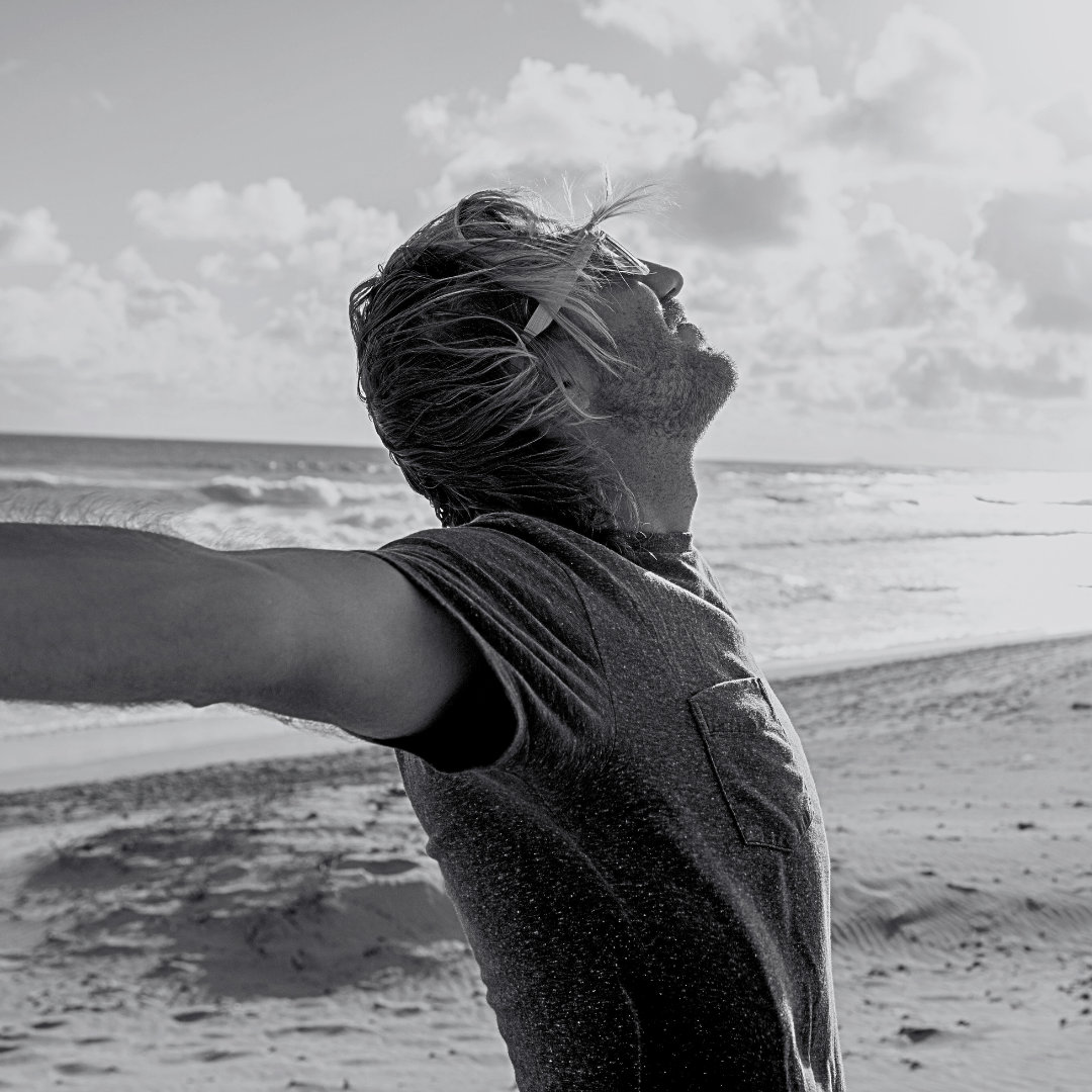 Man with long hair at the beach with arms outstretched, enjoying the breeze, black and white photo.