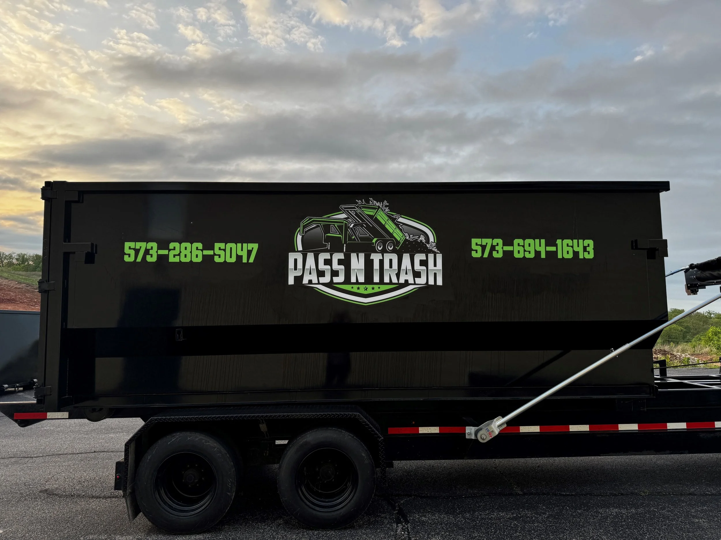 A black trash removal trailer with the logo 'Pass N Trash' and phone numbers 573-286-5047 and 573-694-1643, parked outdoors during sunset.