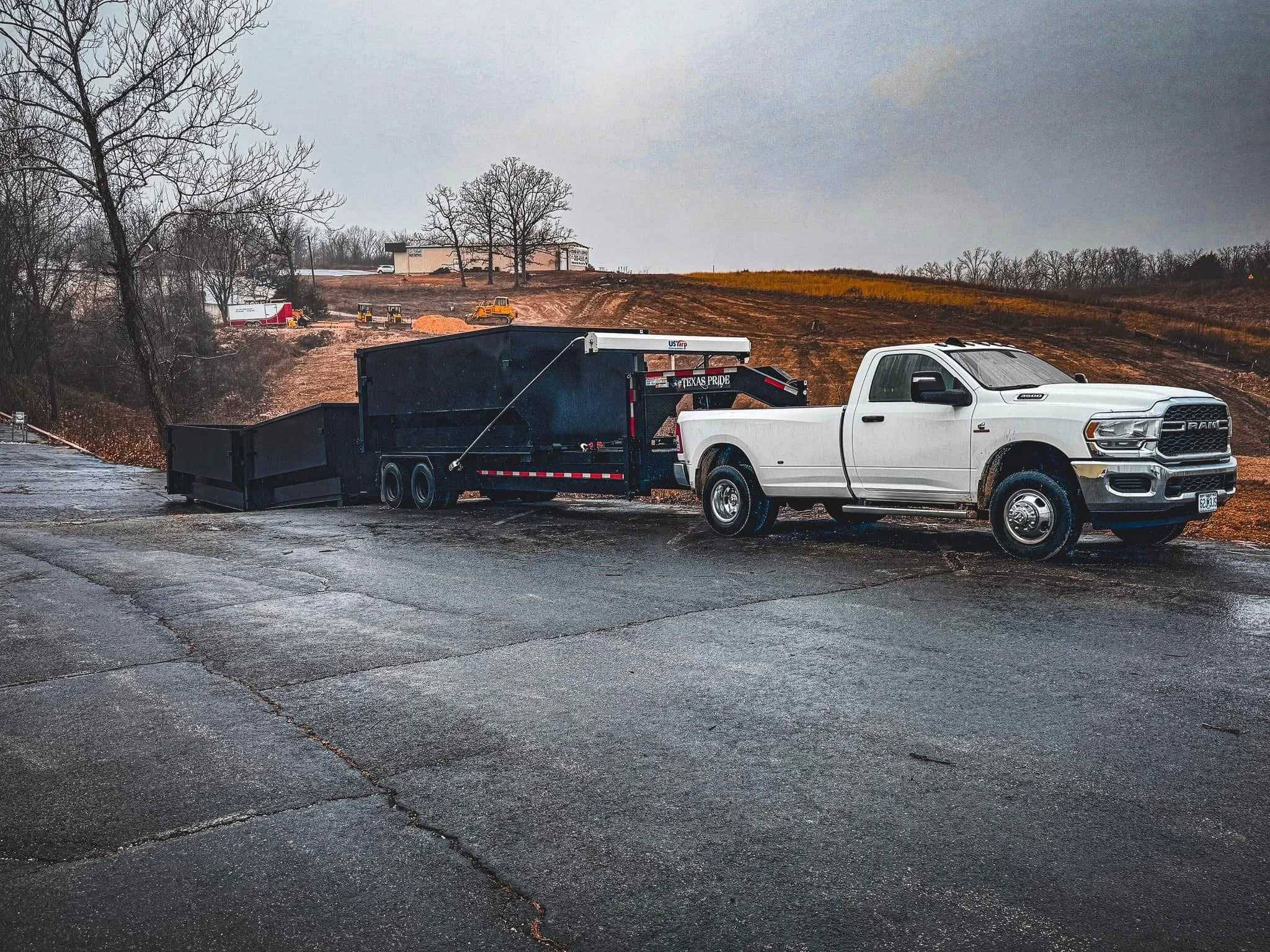 A white Ram pickup truck towing a black utility trailer on a wet asphalt road, with bare trees and a construction site in the background under a gray, cloudy sky.