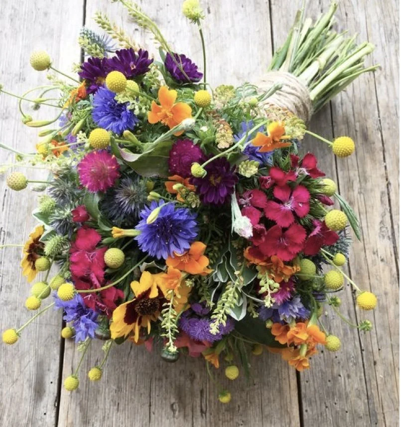 Colorful bouquet of mixed flowers including purple, pink, yellow, orange, and blue blooms with green foliage, resting on a wooden surface.