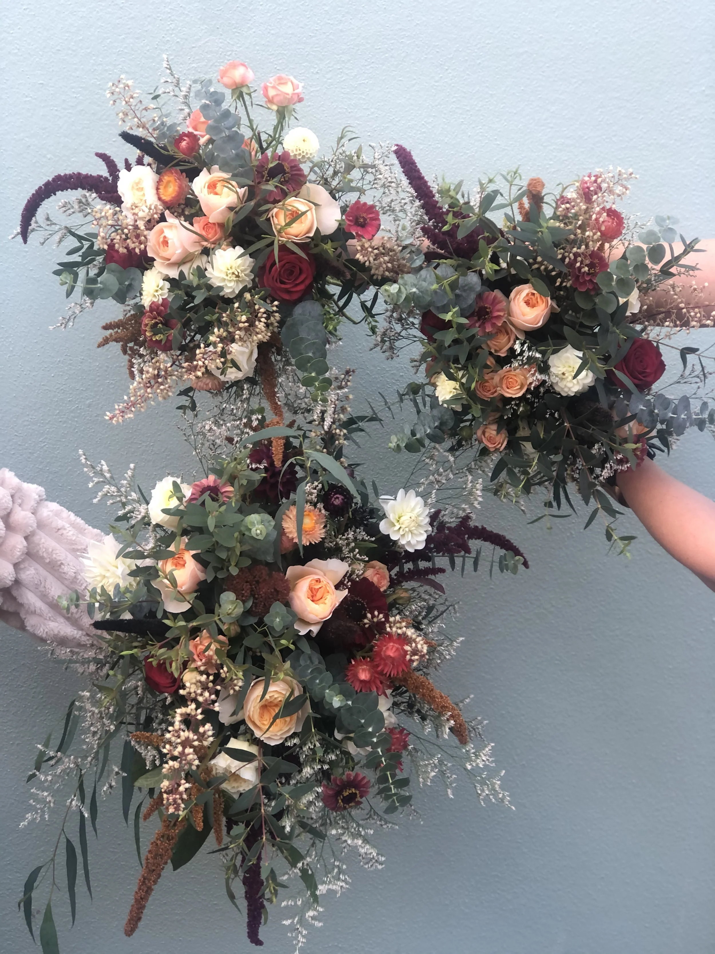 A long cascading floral arrangement with roses, dahlias, eucalyptus, and other greenery against a light blue background.