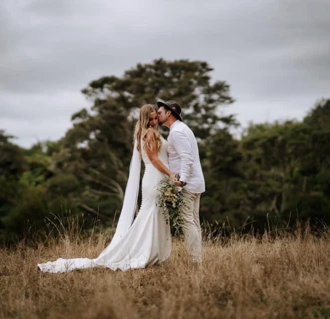A bride and groom standing in a grassy field, facing each other and about to kiss, with trees and an overcast sky in the background. The bride is in a white wedding dress holding a bouquet, and the groom is dressed in light-colored attire with a hat.