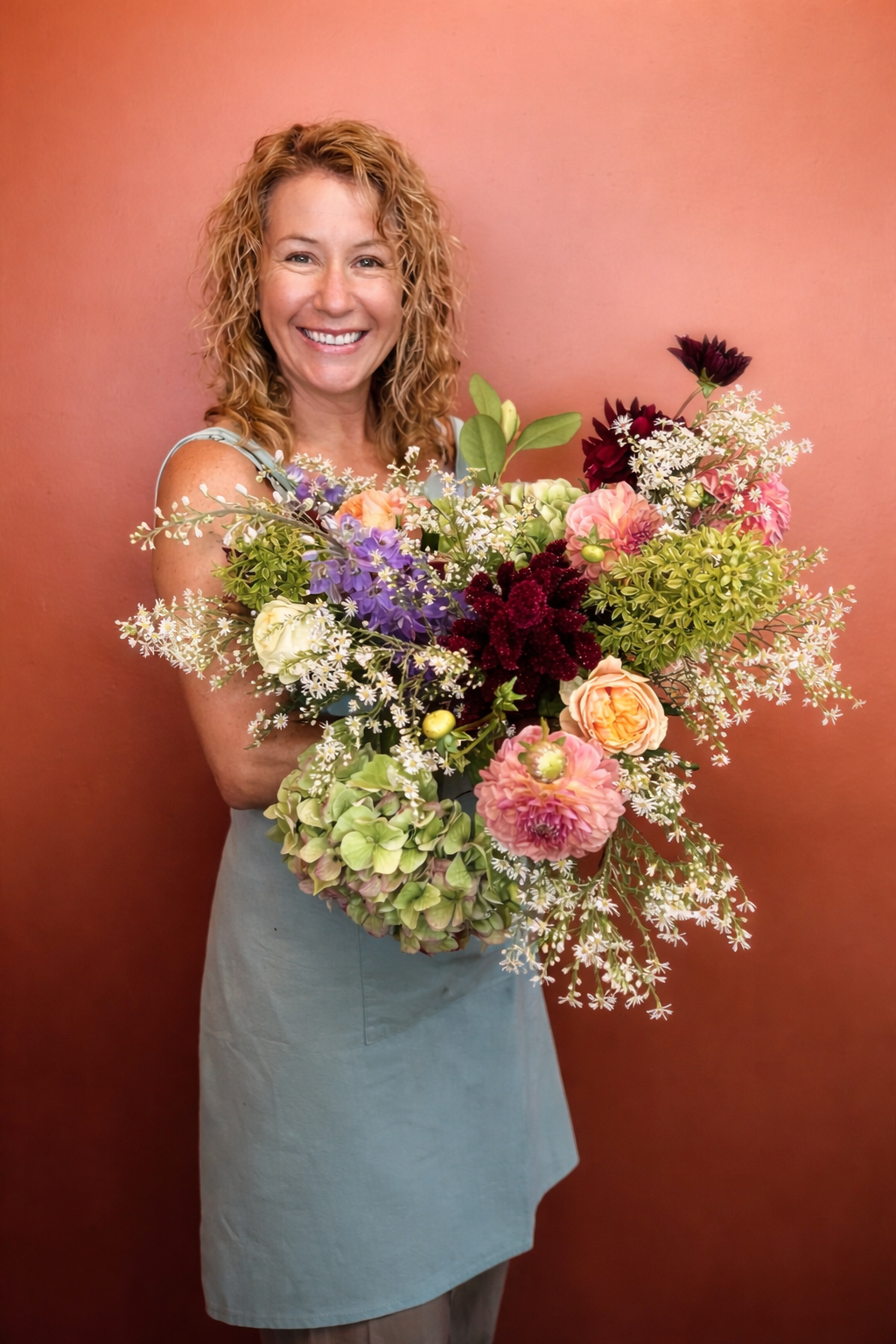 A woman with curly blonde hair smiling while holding a large bouquet of colorful flowers against a pink wall.