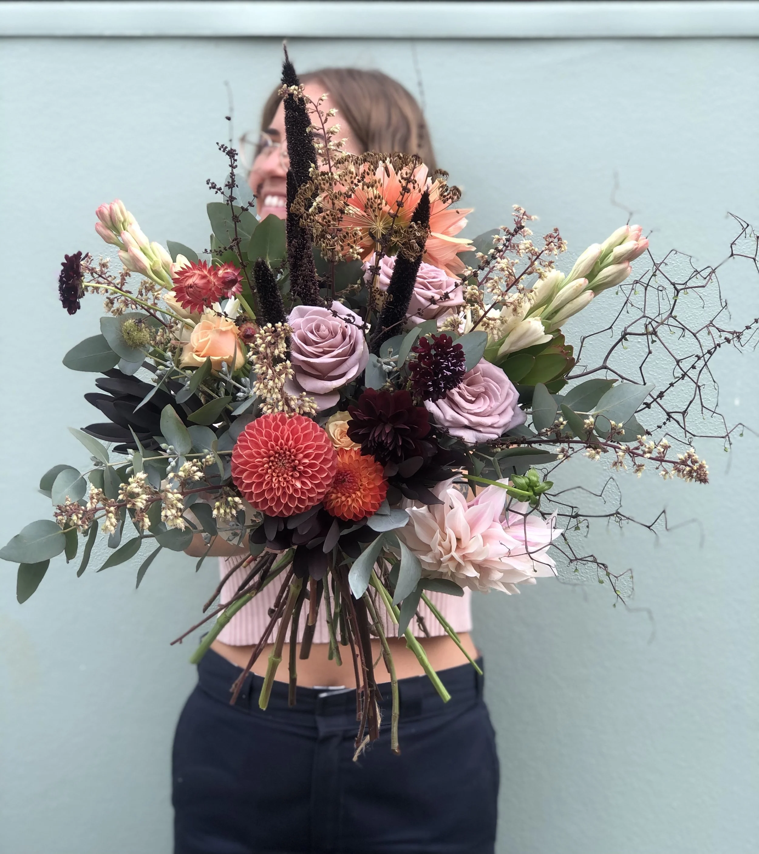 A person holding a large bouquet of multicolored flowers in front of their face, with a light blue wall in the background.