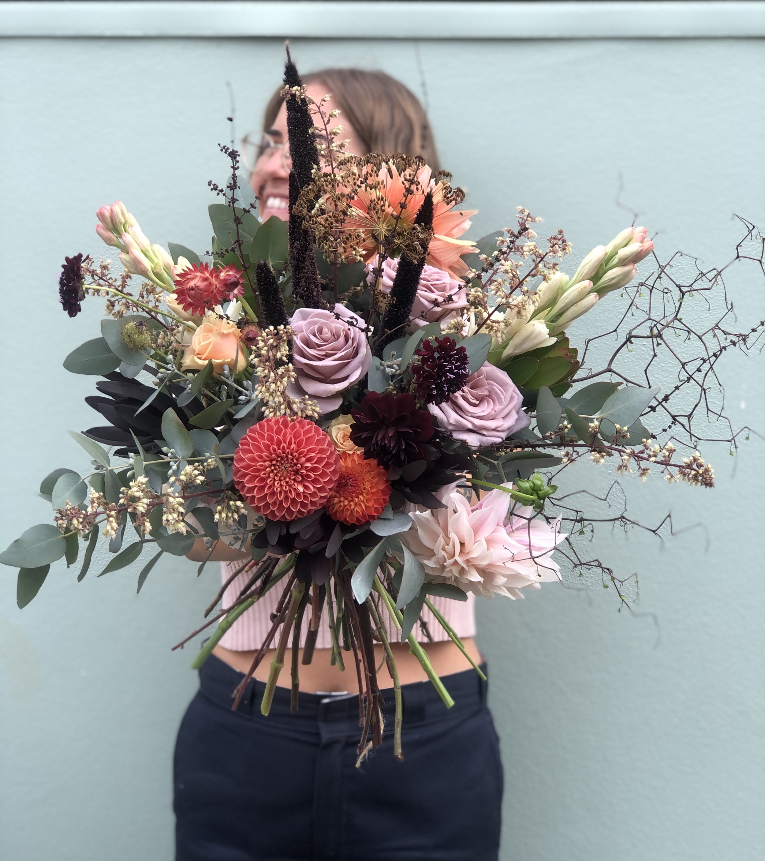 Person holding a large bouquet of assorted flowers in front of their face, standing against a light green wall. For flower deliveries within Central Otago and surrounding areas.