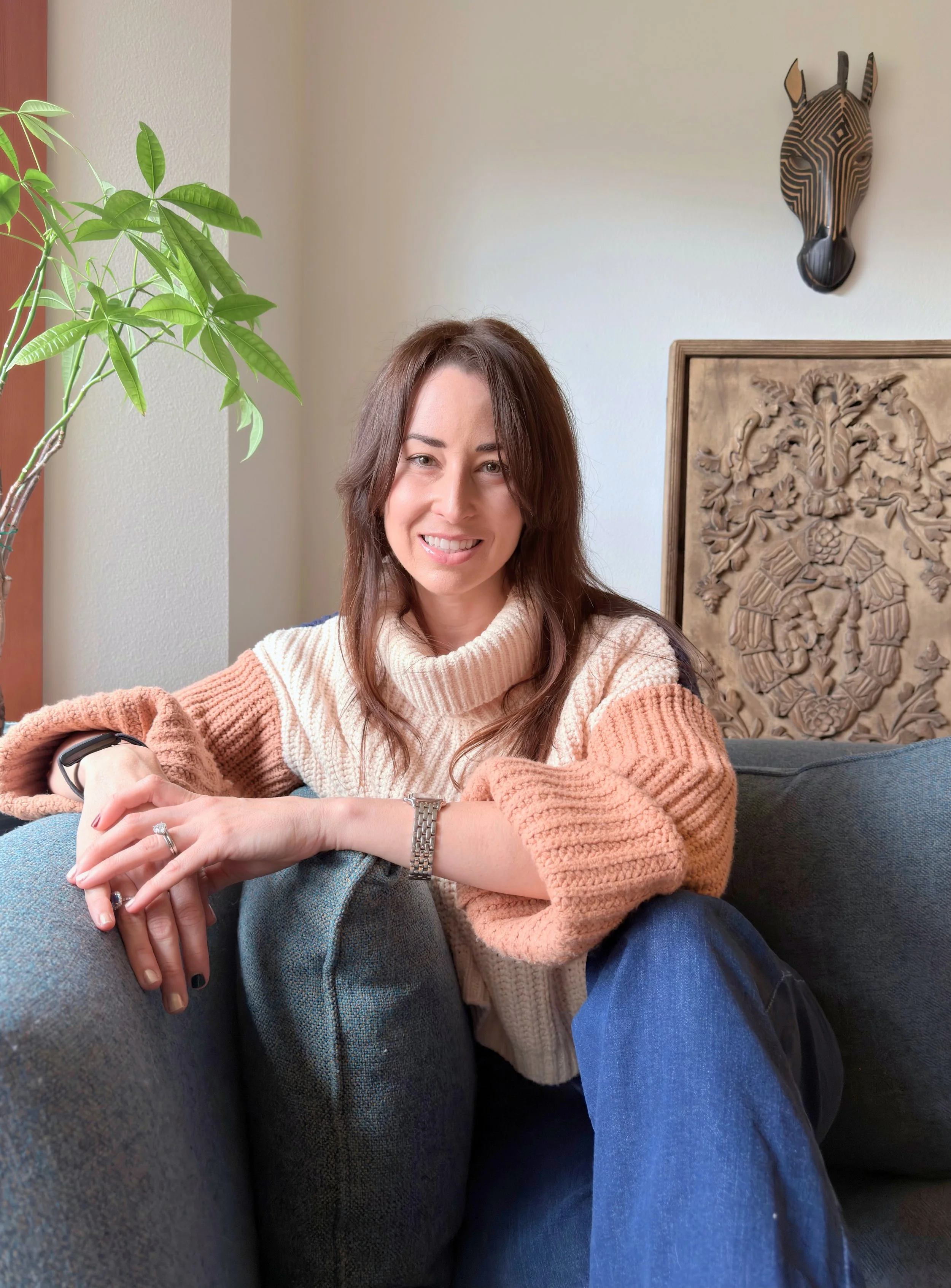 Smiling woman sitting on a dark green couch in a cozy room with a large plant, a decorative carved wood panel, and a zebra mask wall hanging in the background.