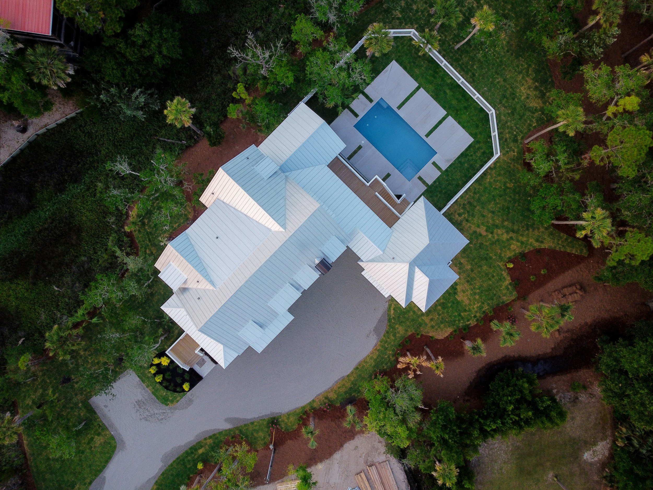 Aerial view of a house with a blue swimming pool and surrounding green lawn, trees, and a driveway.