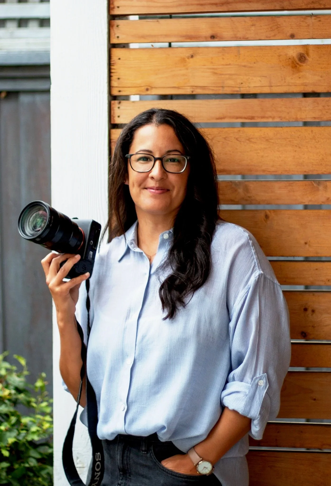 A woman with dark hair, glasses, and a watch, holding a camera, standing outdoors next to a wooden fence.