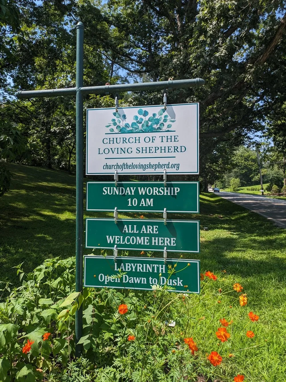 Sign for the Church of the Loving Shepherd with information about Sunday worship at 10 am, welcoming everyone, and a labyrinth open from dawn to dusk, set in a grassy area with flowers and trees.