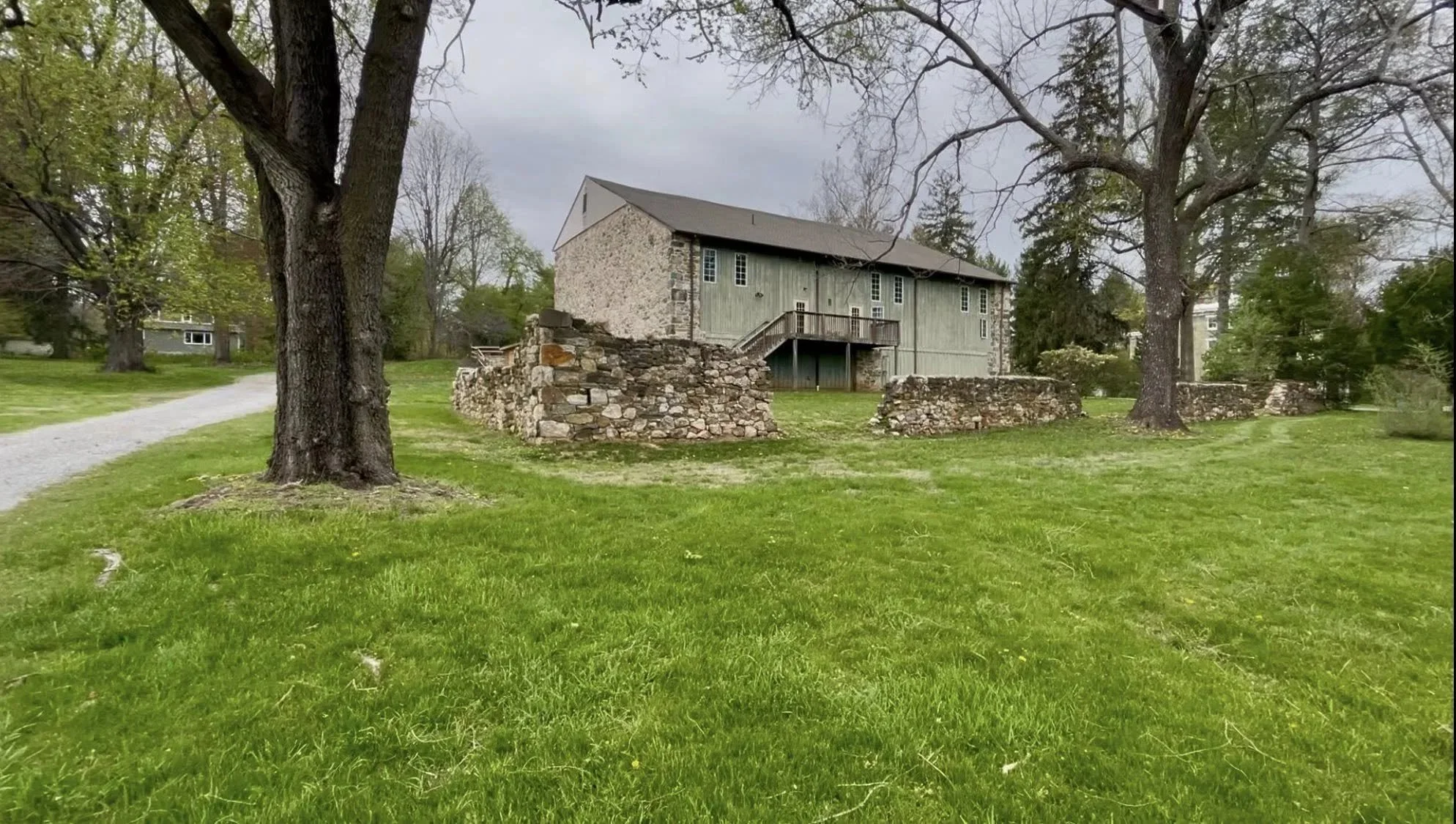An old stone and wood barn with a wooden staircase, surrounded by a grassy lawn and large trees under a cloudy sky.