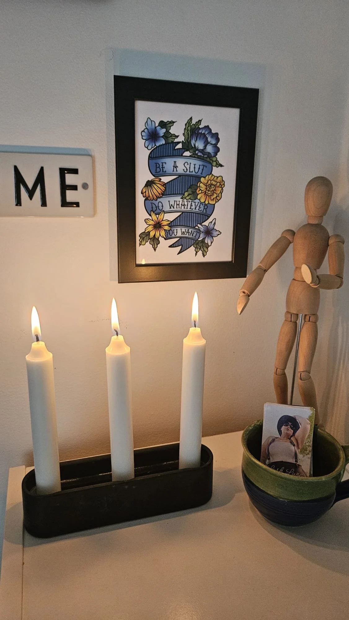 A wall decorated with a framed tattoo-style art print of flowers and the words "Be a Slut, Do Whatever You Want", a wooden mannequin, a candle holder with three lit candles, a black and green ceramic mug with a photo of a woman, and part of a sign wi