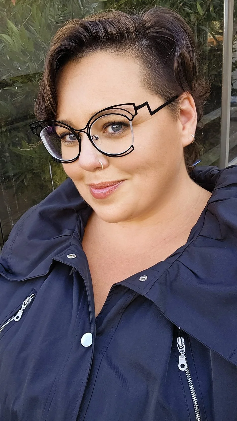 A woman with short, dark, styled hair and glasses with a unique, artistic design, wearing a navy blue jacket with zippers and buttons, smiling outdoors with greenery in the background.