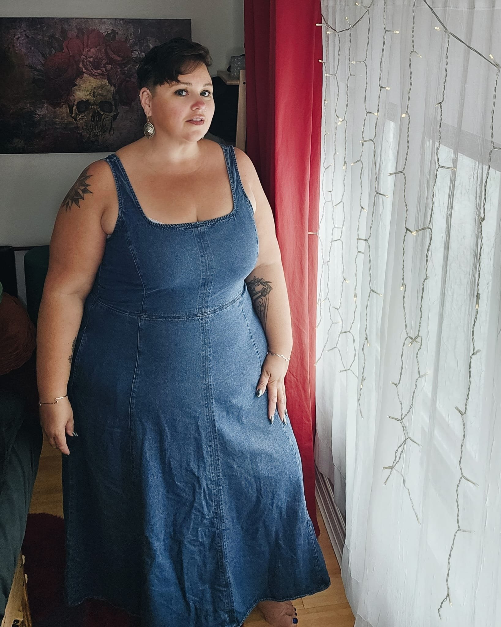 A woman with short dark hair standing indoors near a window with string lights, wearing a sleeveless denim dress and earrings.
