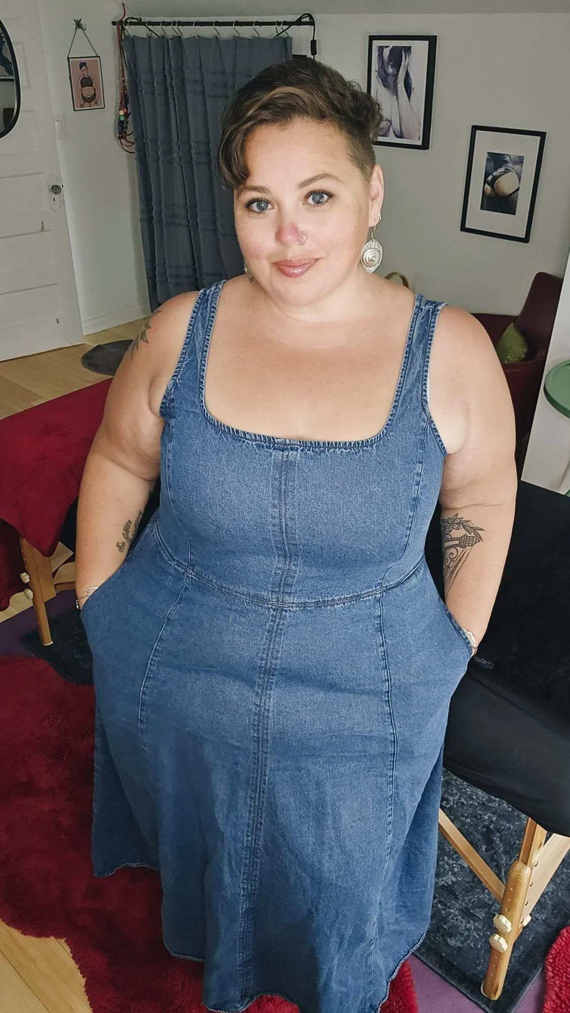 A woman with short, brown hair and blue eyes wearing a sleeveless blue denim dress standing indoors.