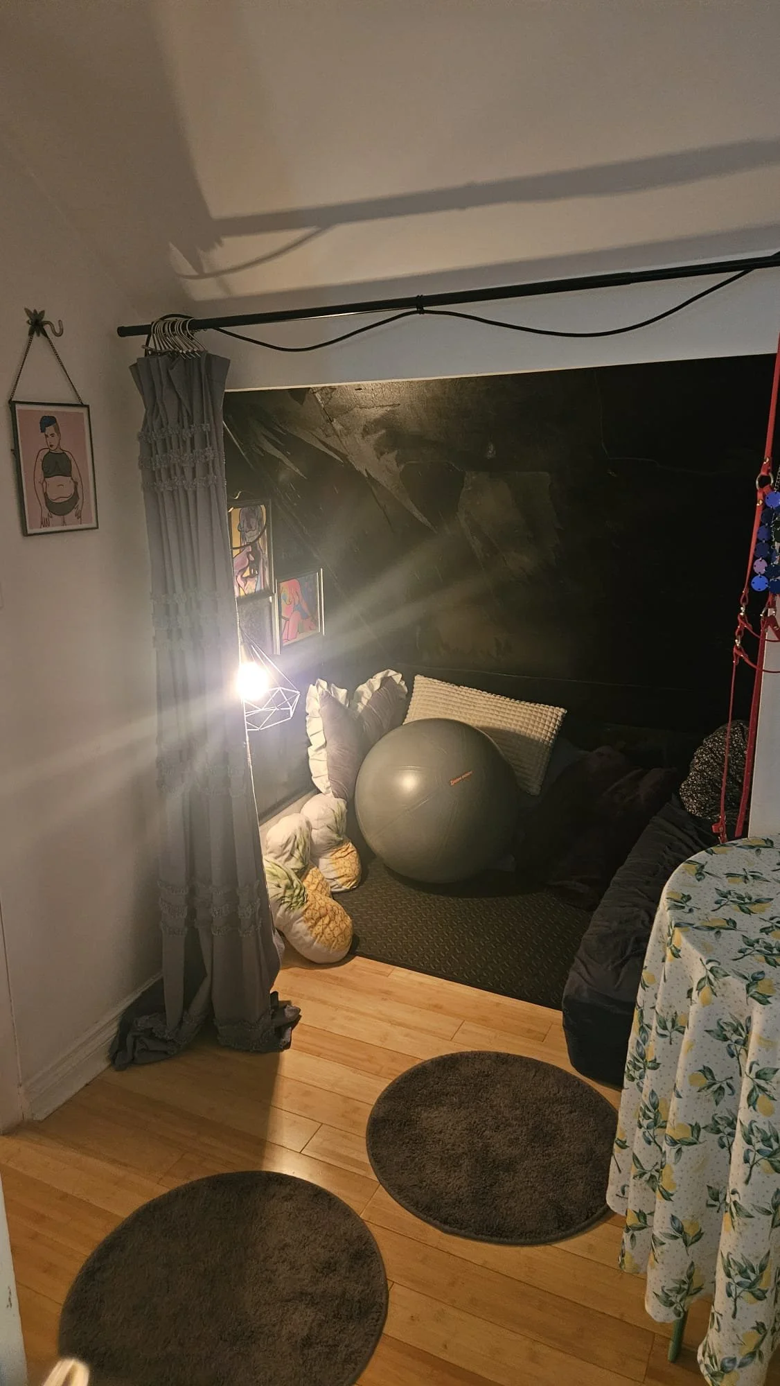 A cozy corner of a room with a low ceiling, black wall, and wooden flooring. It features a bed with pillows, a large exercise ball, framed artwork on the wall, a lamp, and a curtain.