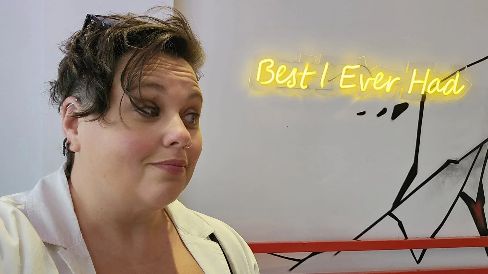 A woman with short curly hair, earrings, and a small nose ring taking a selfie in front of a wall with a neon sign that reads "Best I Ever, Had". The wall has a black geometric design.