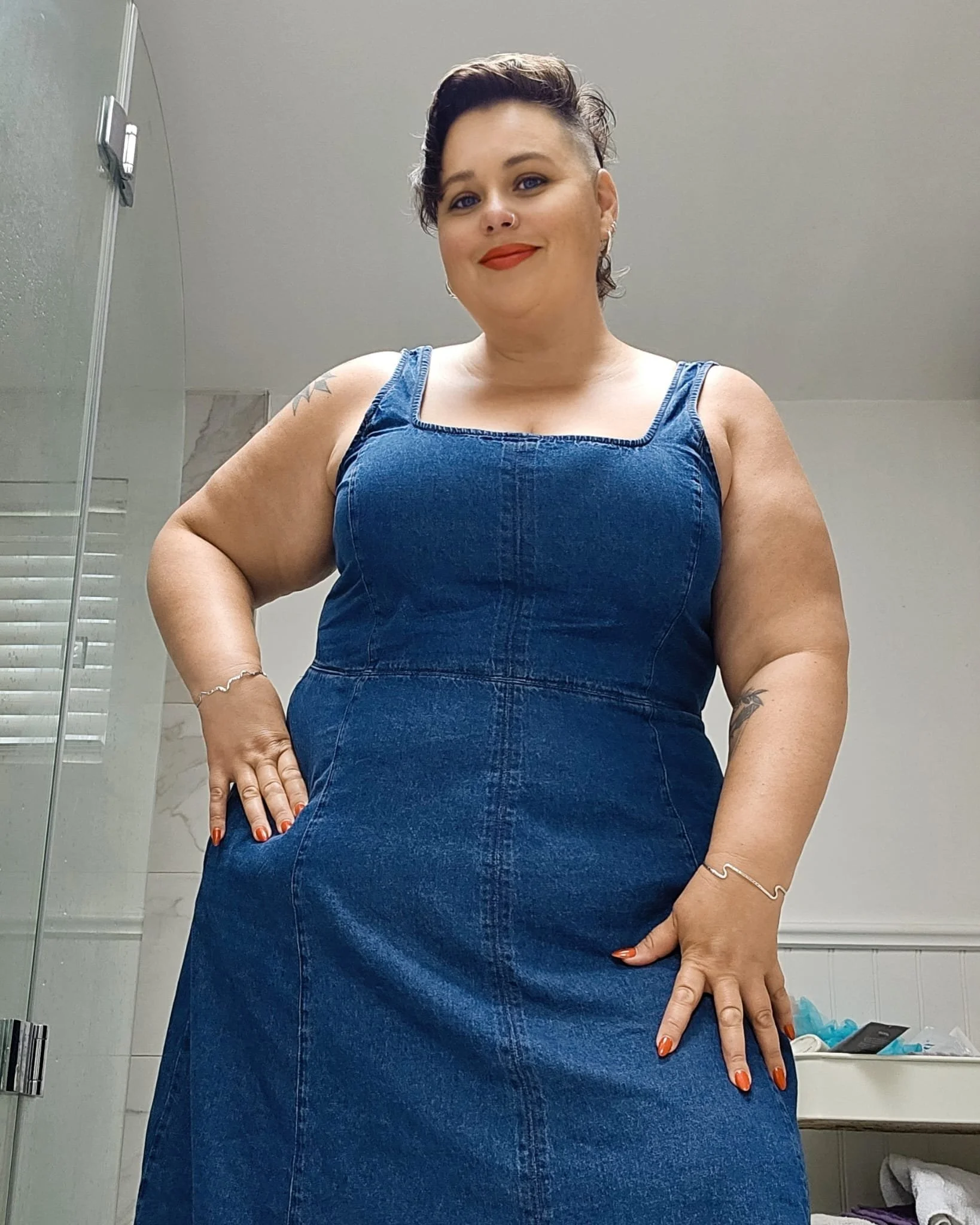 Woman with short curly hair wearing a sleeveless denim dress in a bathroom.