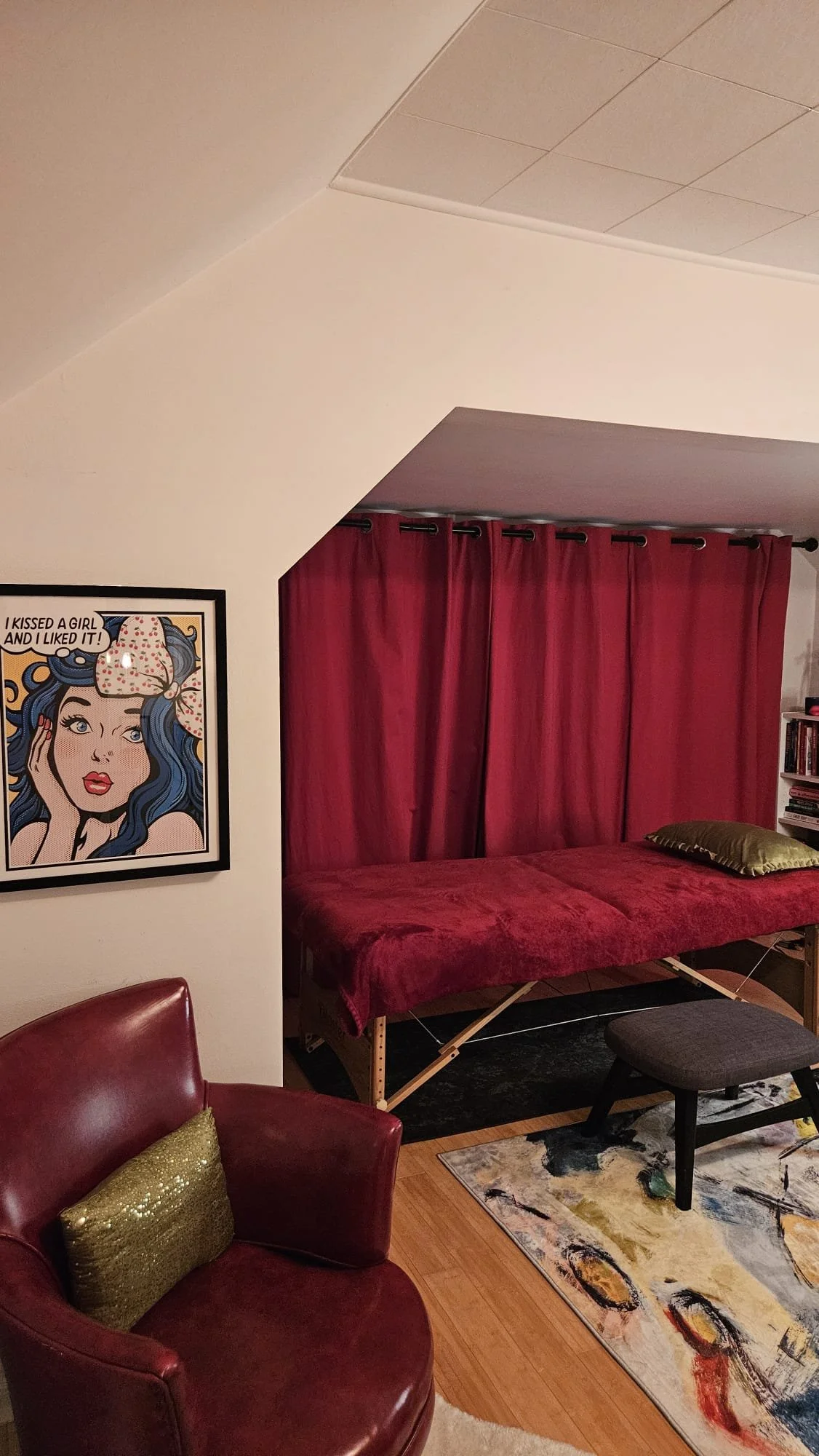 A room with a red velvet armchair with a gold cushion, a colorful abstract rug, a bed with a red velvet canopy or curtains, a picture of a woman with blue hair and a speech bubble saying "I kissed a girl and I liked it!", and a bookshelf with books.