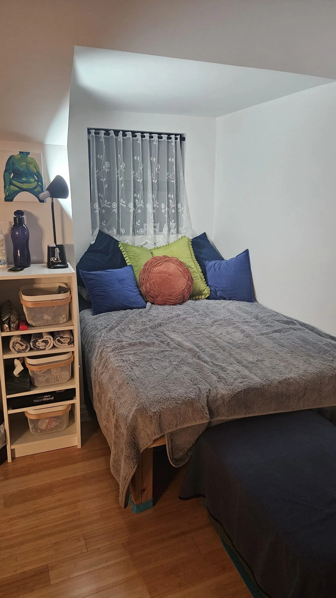 A cozy bedroom with a bed covered in a gray blanket, decorated with colorful pillows, a small window with lace curtains, a nightstand with a lamp, a black water bottle, and a shelf with storage bins and rolled towels.