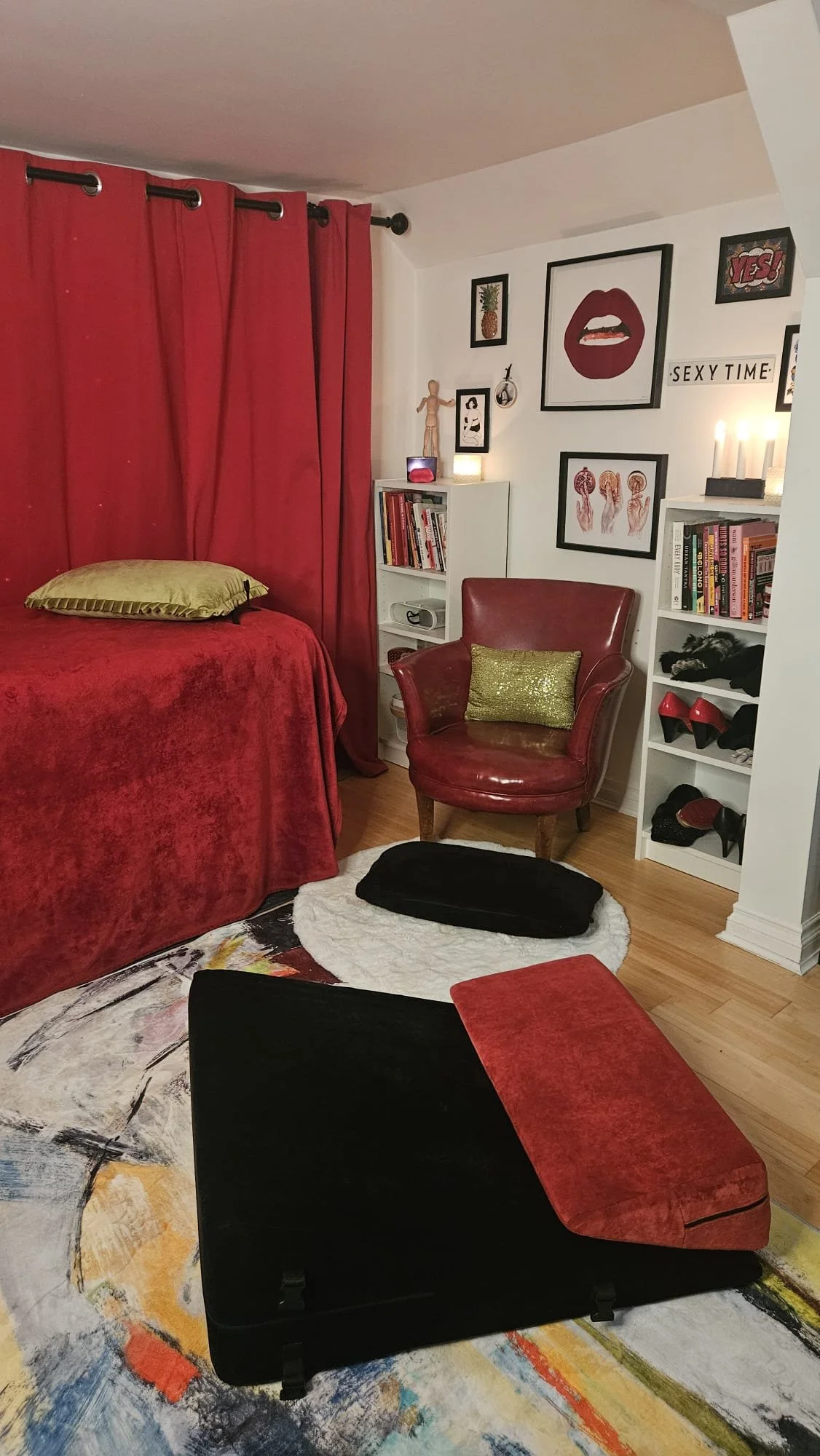 A cozy bedroom corner with a bed covered in red blanket and a gold pillow, a red armchair with a gold throw pillow, a white rug, colorful abstract rug on hardwood floor, and wall art including a large lips print, framed pictures, candles, shelves wit