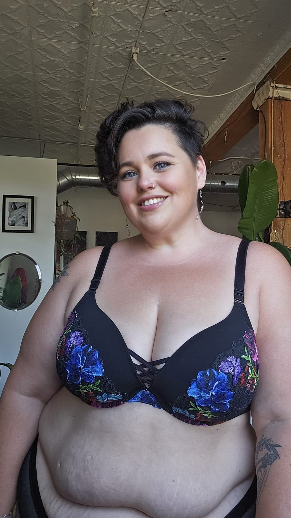 A woman with short dark hair and blue eyes smiling, wearing a black floral bra, standing indoors with a plant and framed pictures in the background.