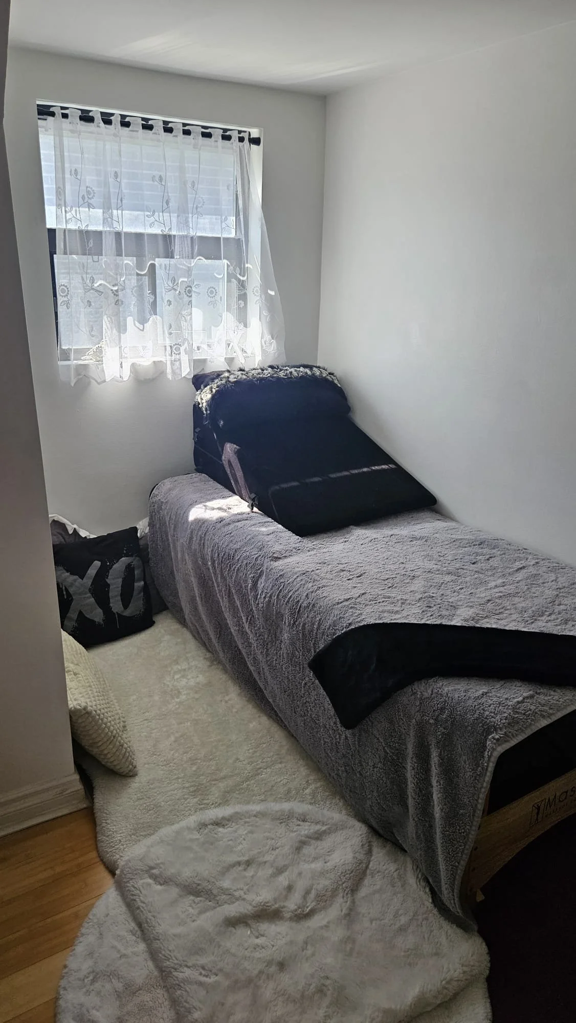 Small bedroom with a single bed covered in gray and black blankets, a window with white floral curtains, a black tote bag with white letters on the floor, and a fluffy white rug.