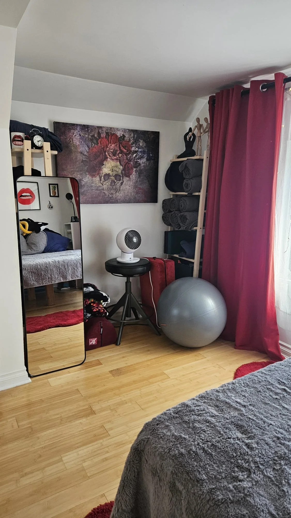 A corner of a bedroom with a mirror, a painting of a skull with flowers, a fan on a stool, a large exercise ball, and shelves with rolled towels. The window has red curtains, and the bed has a gray fuzzy blanket.