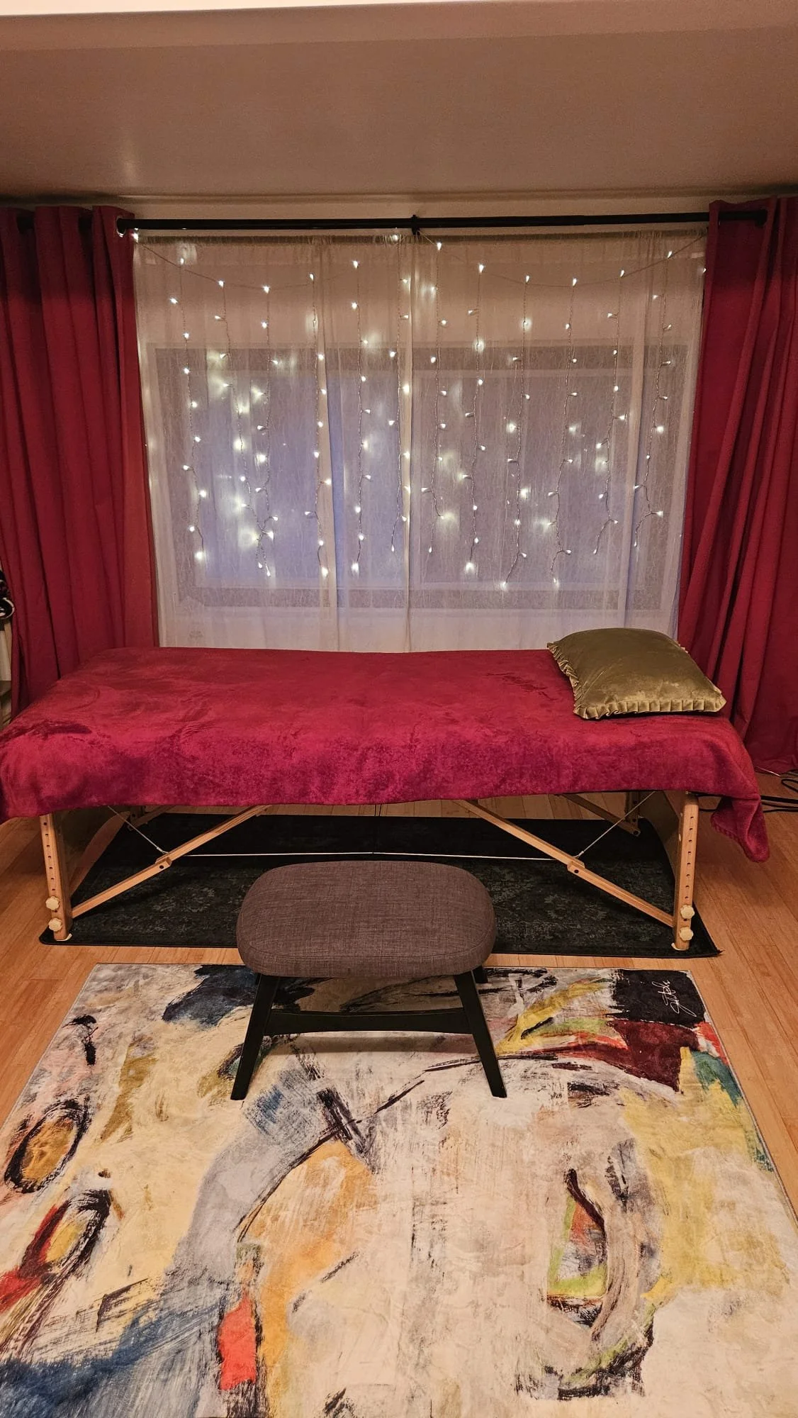 A massage table covered with a red sheet, a brown cushioned stool in front, a framed window with sheer curtains, string lights overhead, and red curtains on either side.