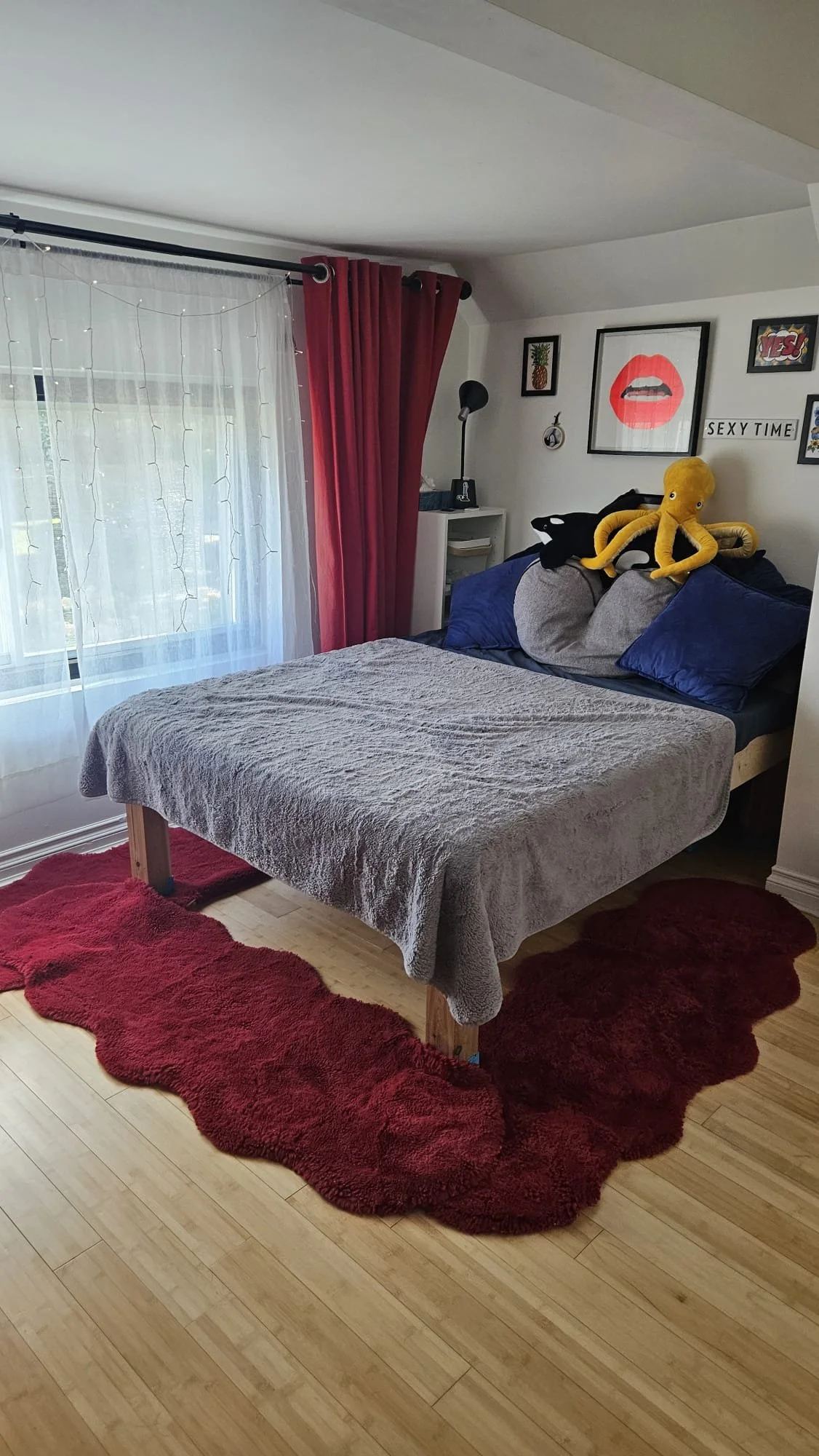 A bedroom with a bed covered by a gray blanket, framed pictures and art on the wall, plush toys on the pillows, red curtains, sheer white curtains, and a red shaggy rug on light wood floor.