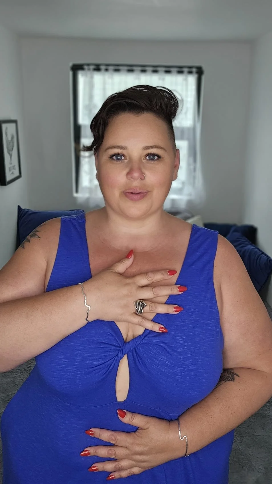 A woman with short dark hair and fair skin, wearing a blue dress with a keyhole cutout, posing indoors with one hand on her chest and the other on her stomach.