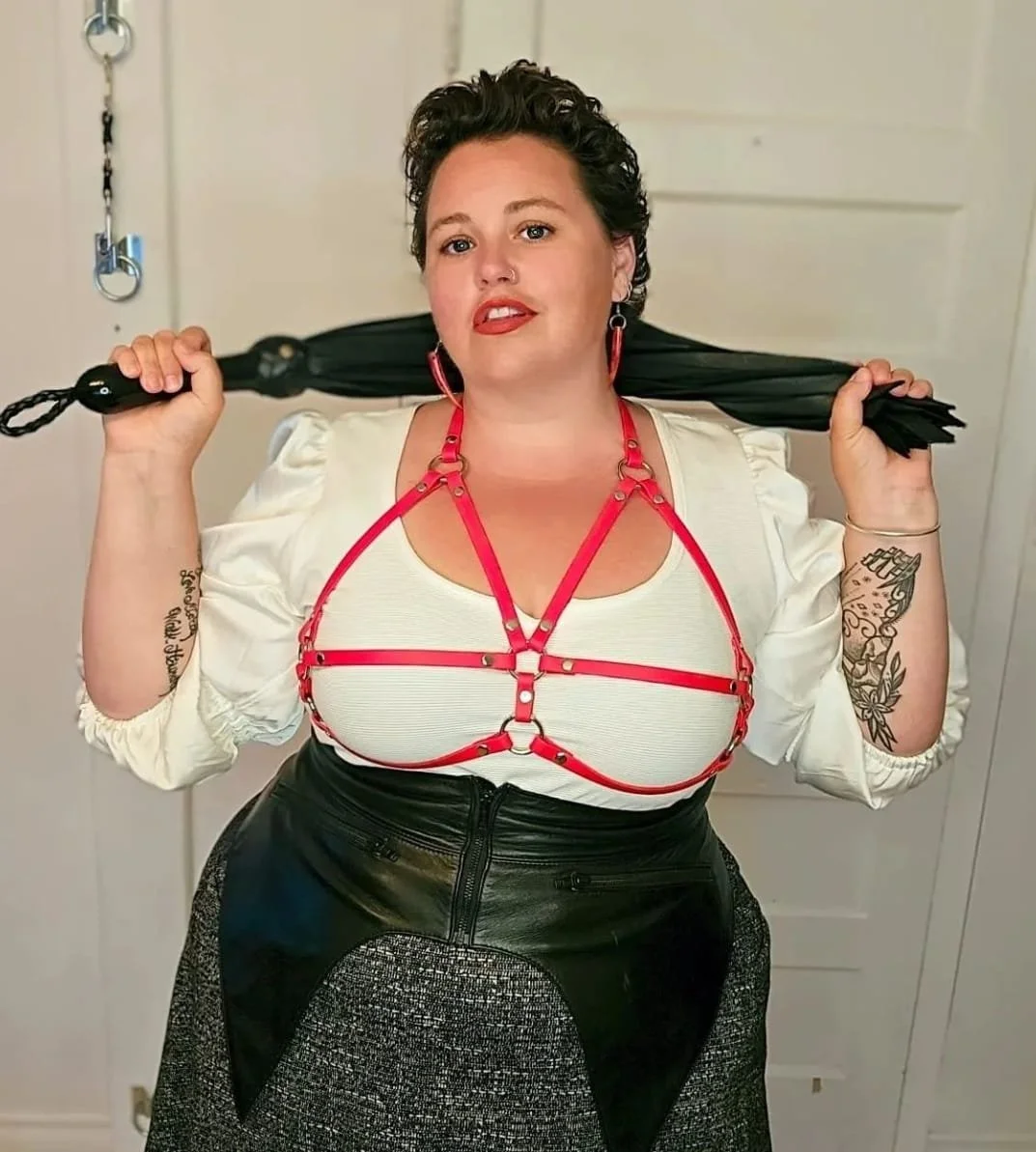 A woman with short curly dark hair, tattoos on her arms, and red lipstick, is holding a black umbrella behind her neck. She is wearing a white blouse with puffed sleeves, a red harness-style top, and high-waisted black leather and metallic skirt.