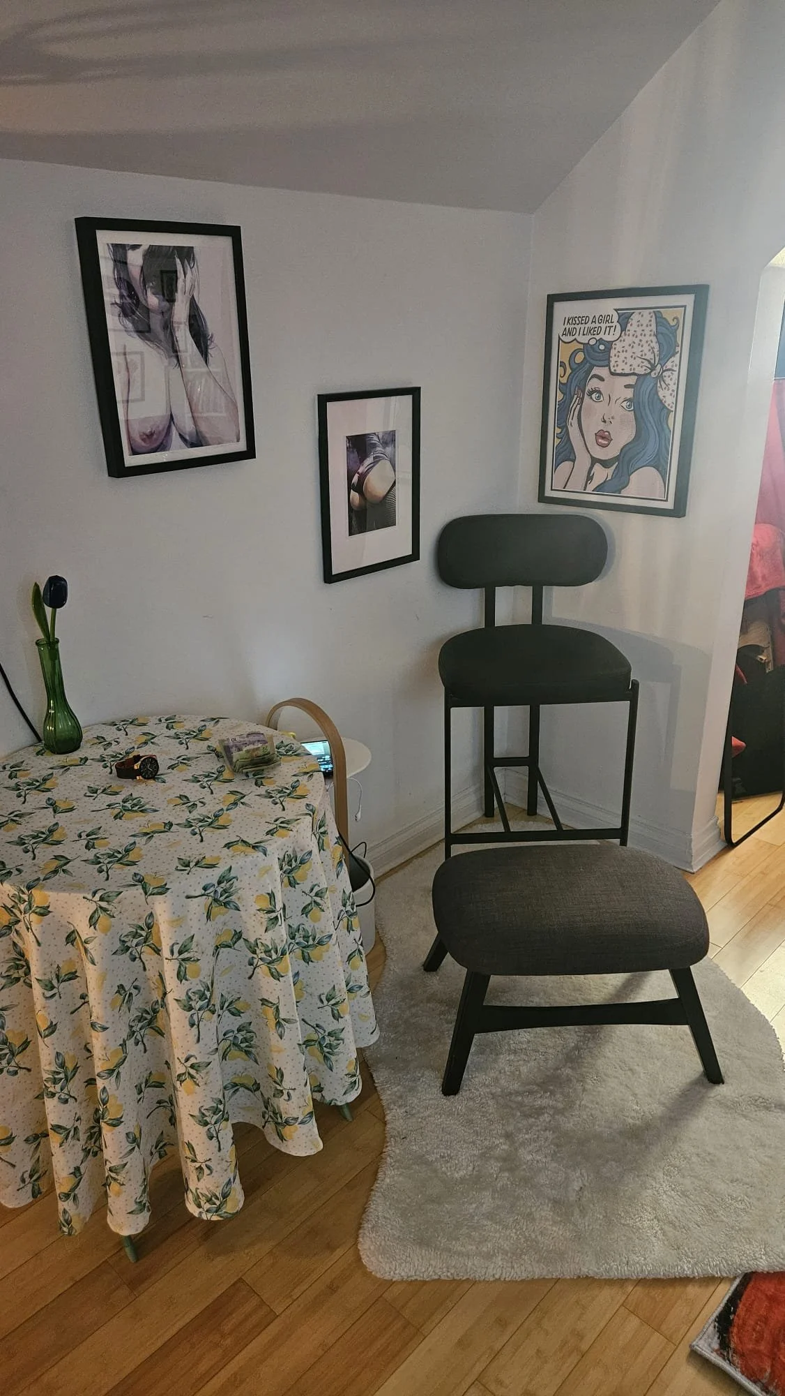 A corner of a room with a round table covered with a floral tablecloth, a green vase, and small objects. There are two black chairs, a white shaggy rug, and several framed art pieces on the white wall, including a pop art portrait of a woman and a co