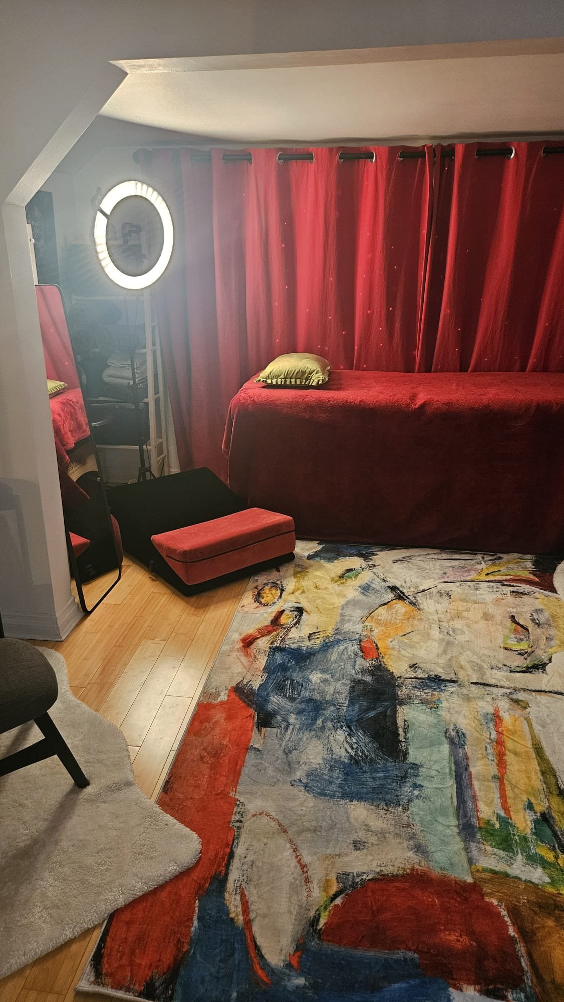 A cozy room with a bed draped in red fabric with a matching curtain, a colorful abstract rug, and a ring light mirror. There are two small stools near the mirror and a chair with a fuzzy white rug on it.