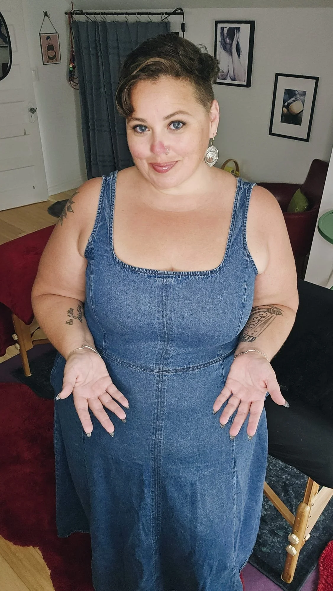 A woman with short hair and earrings wearing a sleeveless denim dress, standing indoors with her hands open, looking at the camera.