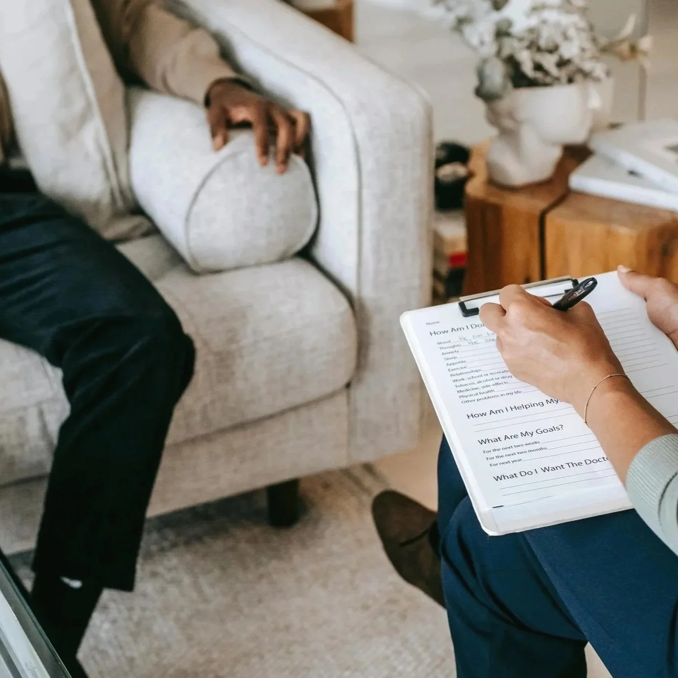 A person holding a clipboard and pen while conducting a mental health assessment with a patient sitting on a sofa.