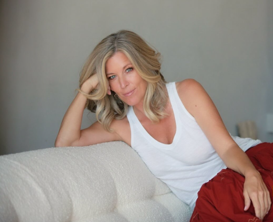A woman with blonde hair, wearing a white tank top, lying on her side on a white couch, resting her head on her hand, and smiling softly.